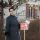 Smiling woman realtor in winter coat standing by a for sale sign with documents, outdoors.